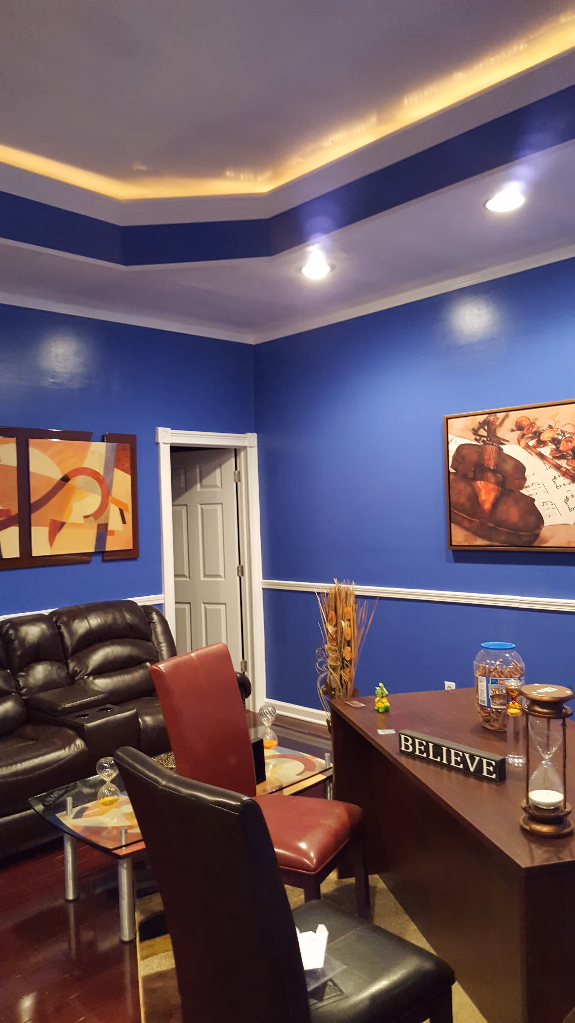 Blue-walled lounge with leather couch, chairs, glass coffee table, and a 'BELIEVE' sign.