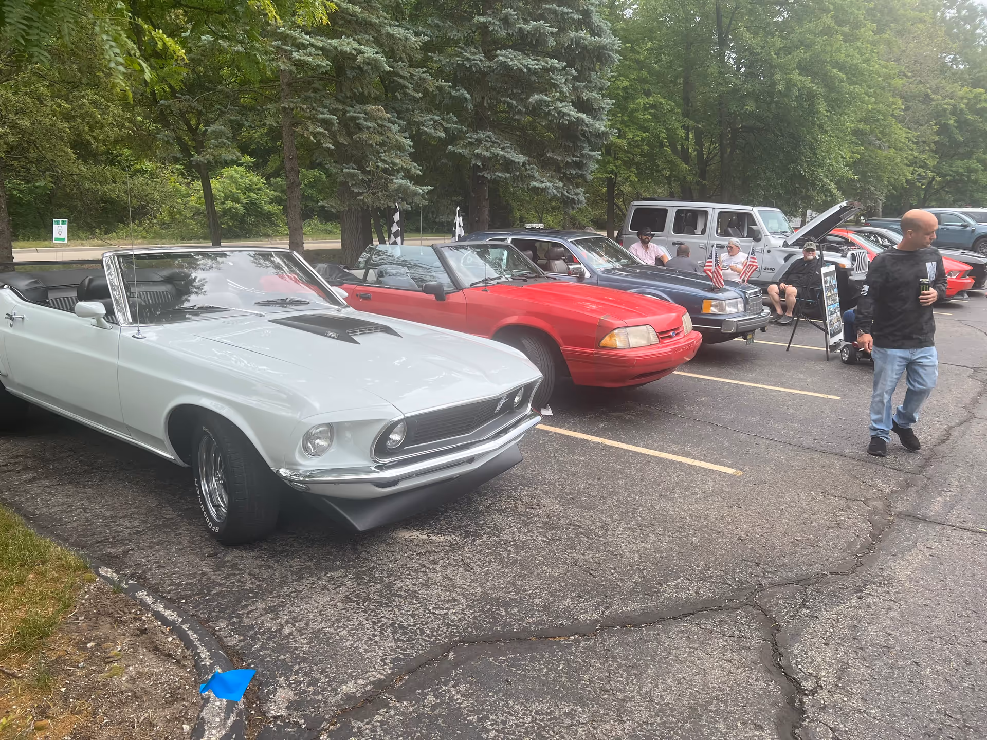A parking lot with several classic and modern cars parked in a row. People are sitting and standing near the cars, some holding drinks. Trees and greenery are visible in the background.