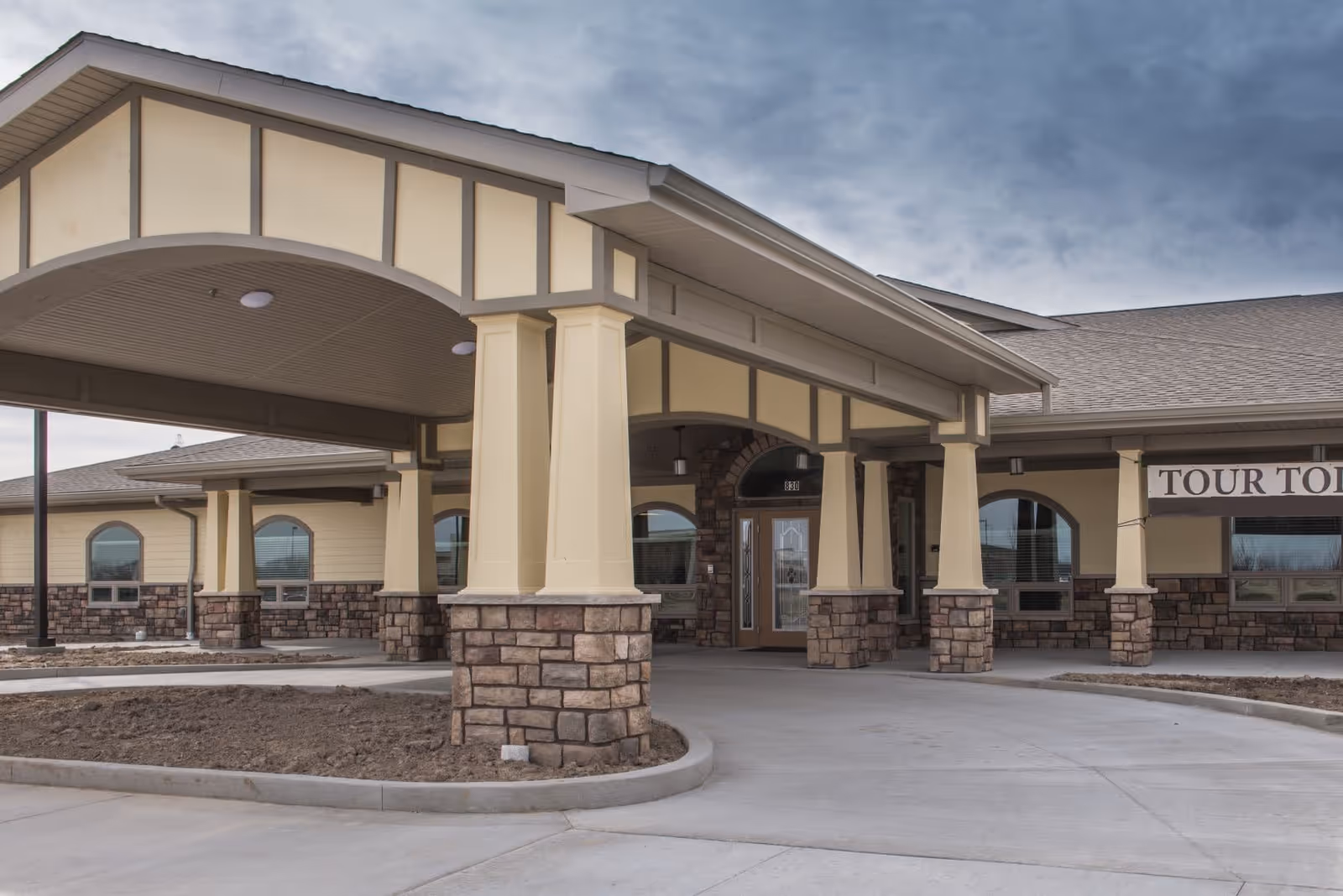 Exterior view of BeeHive Homes Memory Care - Lafayette showing the covered entrance with stone pillars and beige siding under a cloudy sky.