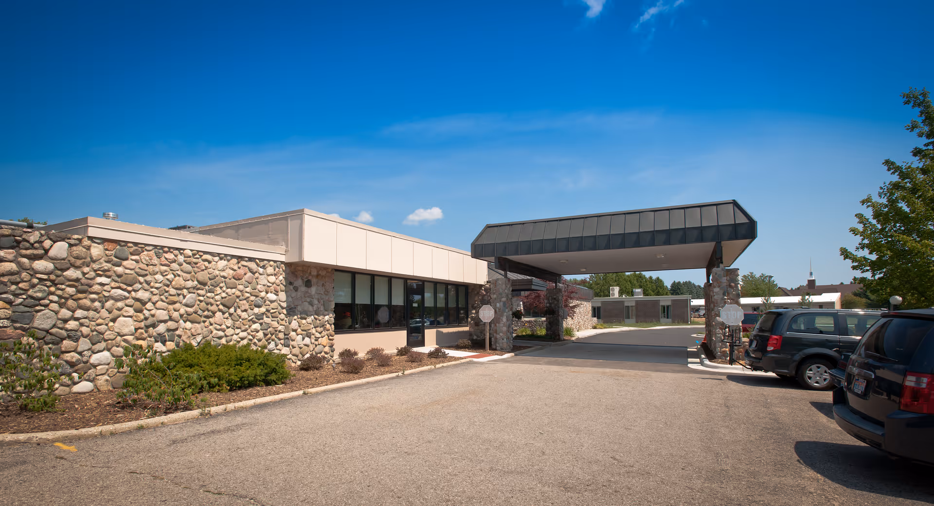 Exterior view of a senior care facility building with stone walls and a covered entrance driveway. Several parked cars are visible on the right side under a clear blue sky.