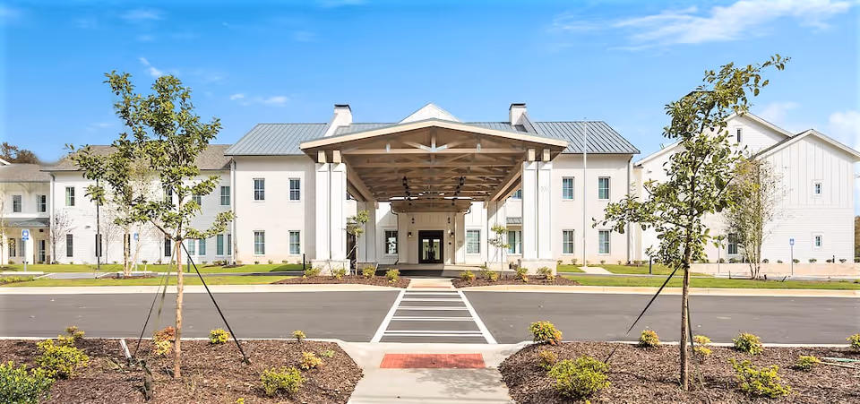 Front exterior view of The Claiborne at Westlake building with a covered entrance, two stories, white walls, and a metal roof. There are young trees and landscaped areas in front, a crosswalk leading to the entrance, and a clear blue sky above.