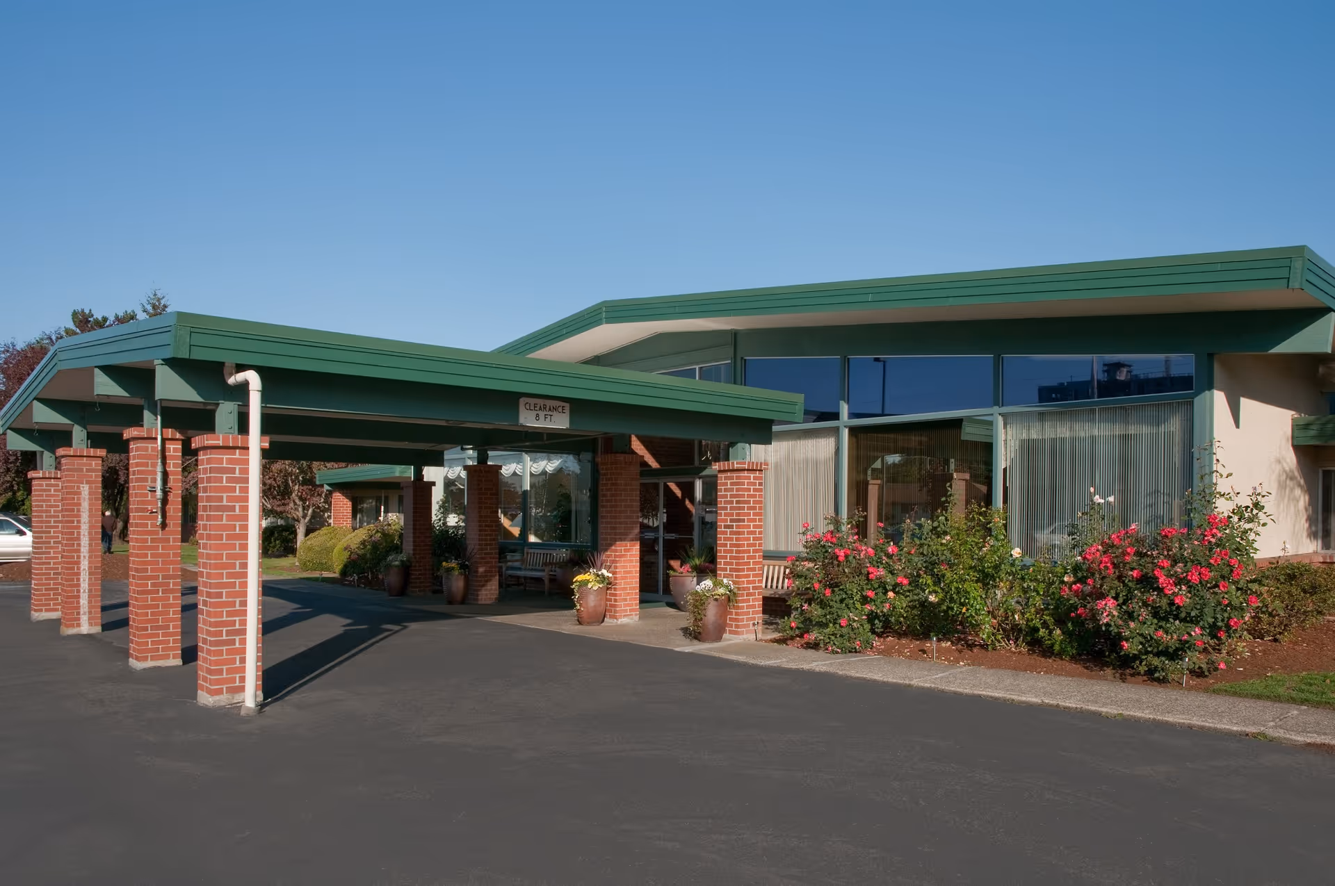Exterior view of the Life Care Center of Puyallup showing the entrance with a green roof supported by brick pillars, a paved driveway, and landscaping with bushes and flowers under a clear blue sky.
