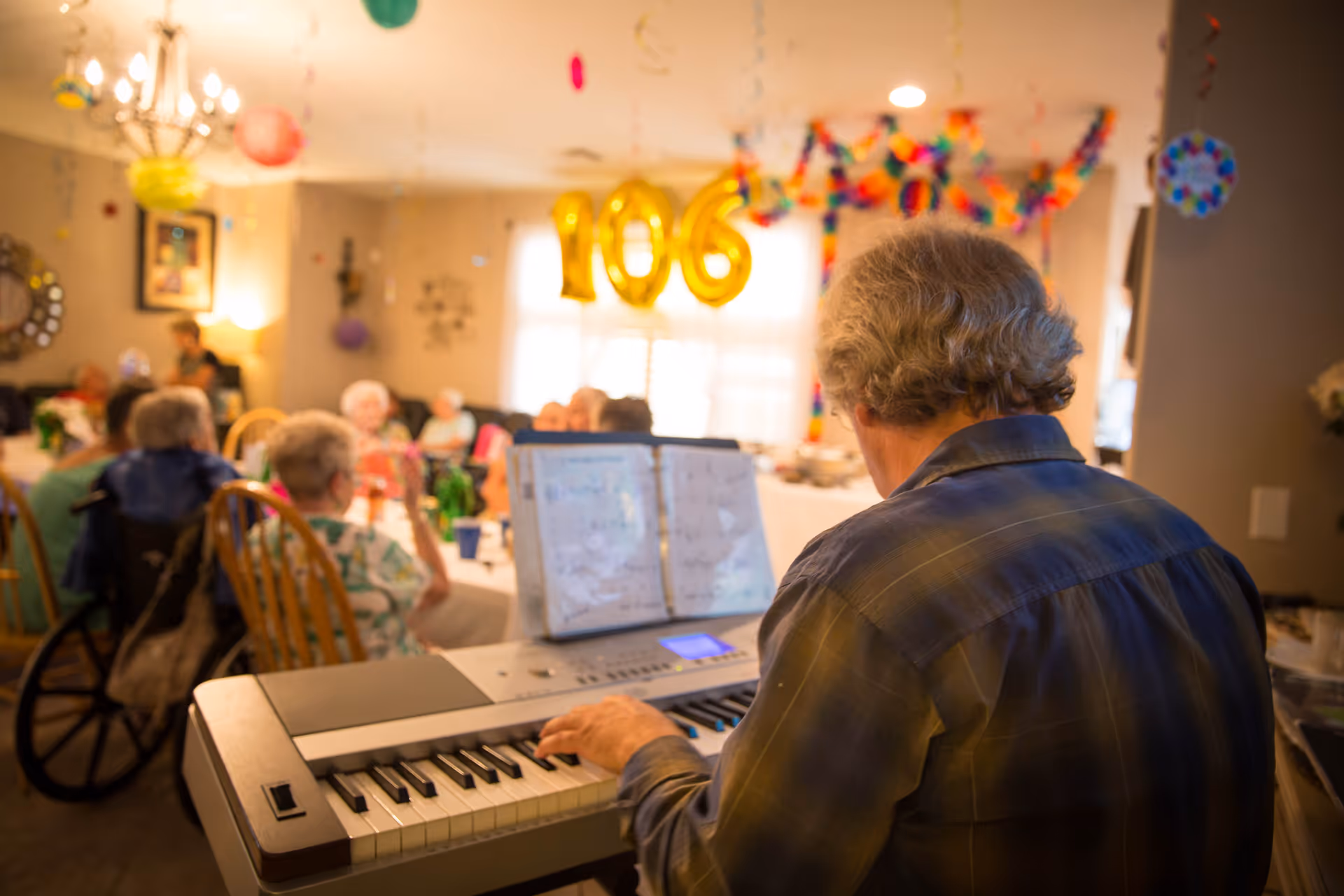 An elderly man playing an electronic keyboard in a decorated room with a group of elderly people seated at tables in the background. Gold balloons forming the number 106 are hanging in the room, indicating a celebration. The room is warmly lit with hanging decorations and a chandelier.
