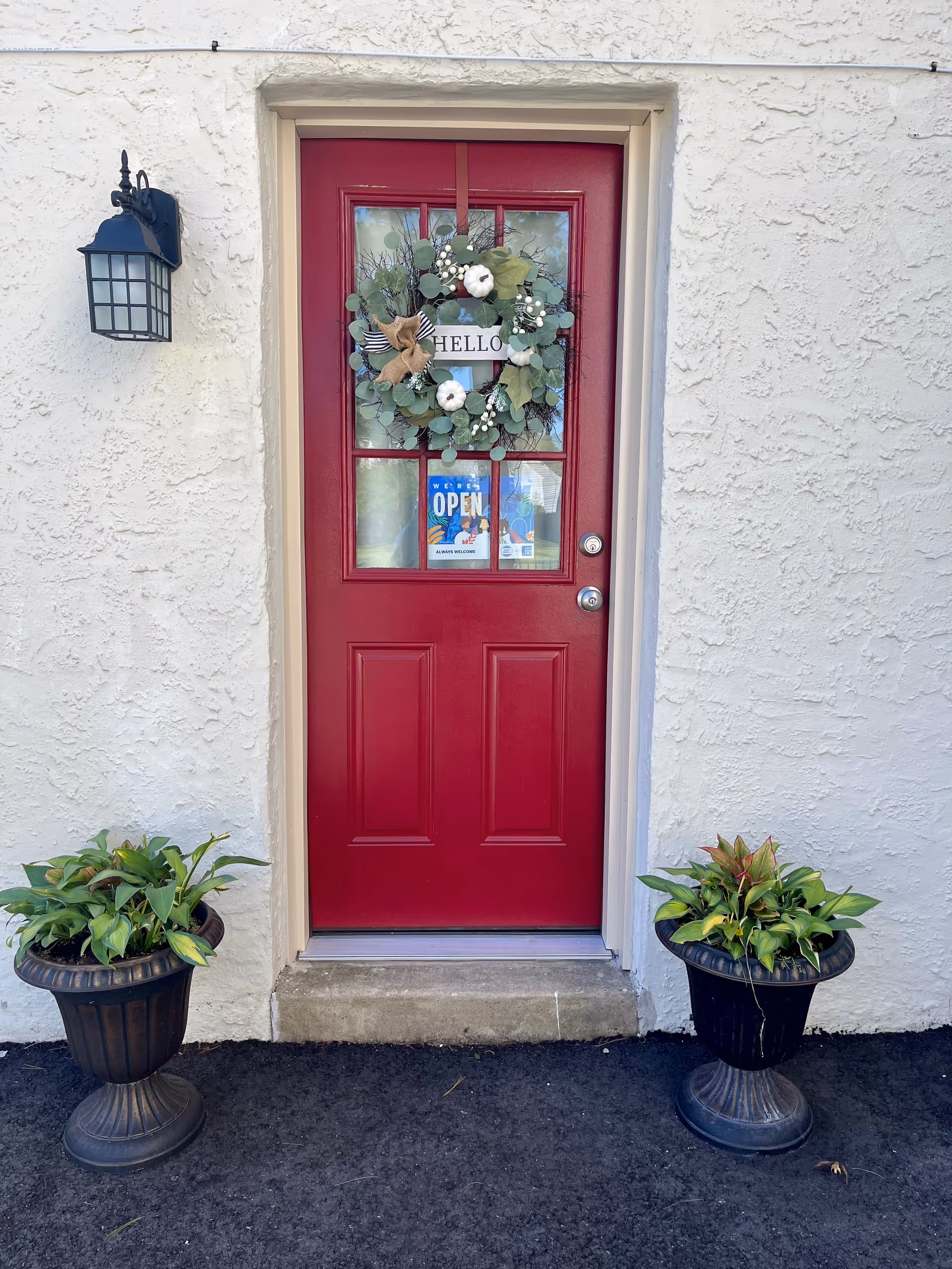 Red front door with wreath, flanked by two potted plants on an exterior wall.
