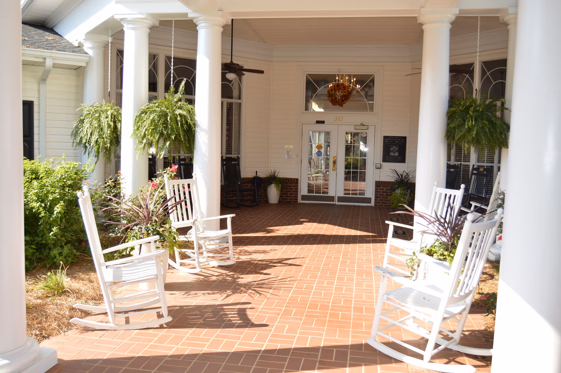 Covered front porch entrance with white columns, hanging ferns and white rocking chairs on a brick floor leading to glass double doors.
