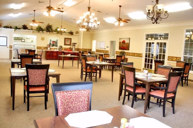 Bright dining room with multiple tables and upholstered chairs, chandeliers, and a buffet/serving area in the background.