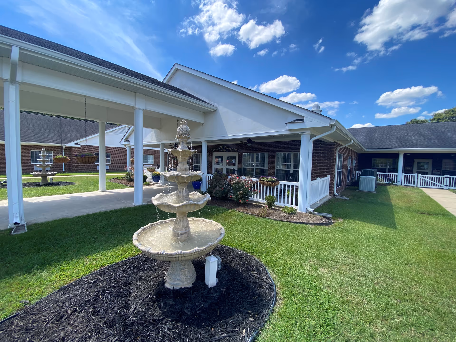 Outdoor view of Liberty Commons Nursing & Rehab Center of Johnston County showing a white multi-tiered fountain in the foreground surrounded by mulch and grass. The building has red brick walls with white columns and railings, and a covered walkway. The sky is blue with scattered clouds.