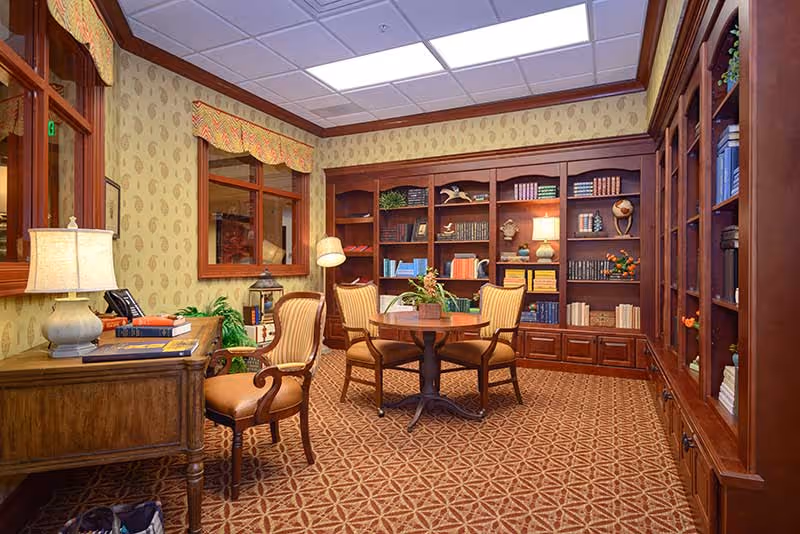 A cozy interior room with patterned carpet and wallpaper, featuring a wooden desk with a lamp and books, a round table surrounded by four upholstered chairs, and large wooden bookshelves filled with books and decorative items. The room has warm lighting and a window with a valance.