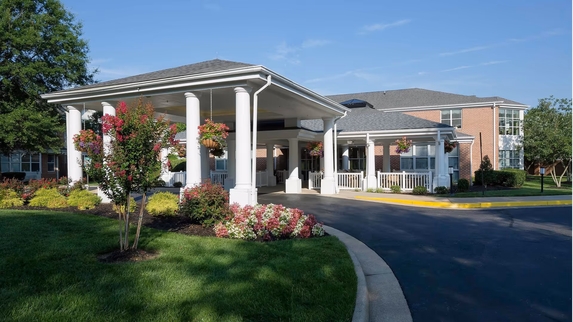 Exterior view of Our Lady of Hope Health Center showing a covered entrance with white columns, well-maintained landscaping with green grass, flowering plants, and trees under a clear blue sky.