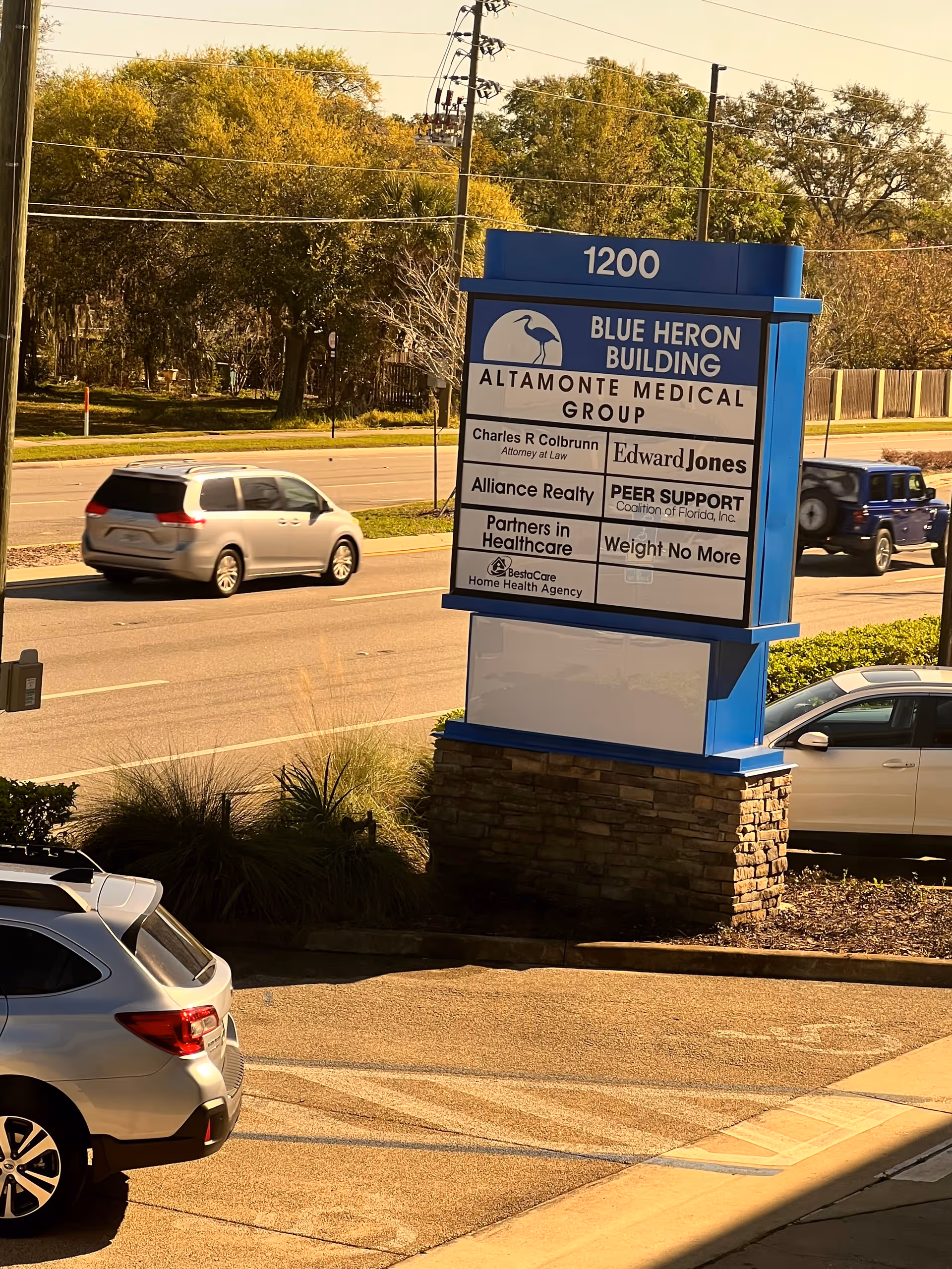 Exterior view of a blue-and-white building sign with tenant listings and nearby cars.