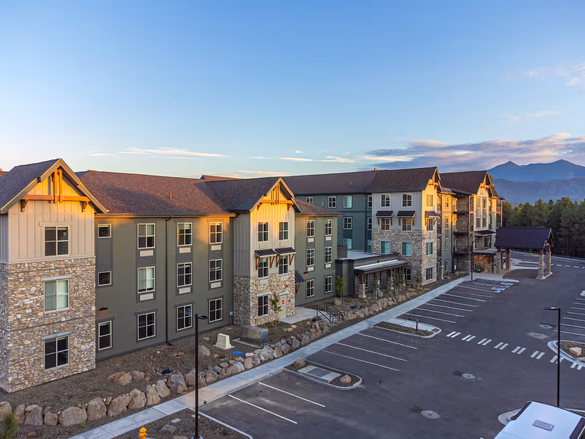 Exterior view of a multi-story senior living building with stone accents, a large parking lot, and mountains in the background.