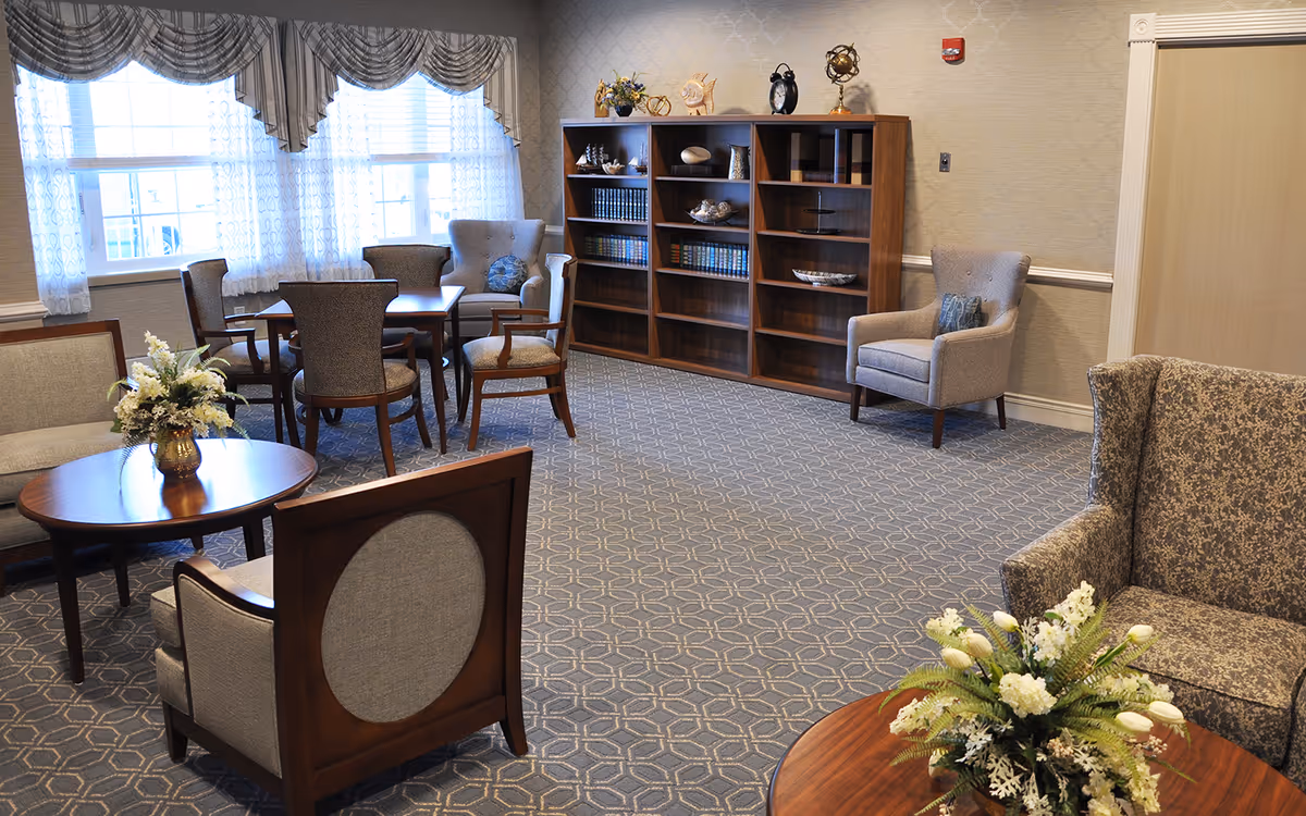 A cozy common area in Nemasket Healthcare Center featuring multiple upholstered chairs and wooden tables arranged on a patterned carpet. There is a large window with sheer curtains allowing natural light to fill the room. A wooden bookshelf with decorative items and books is against the wall.