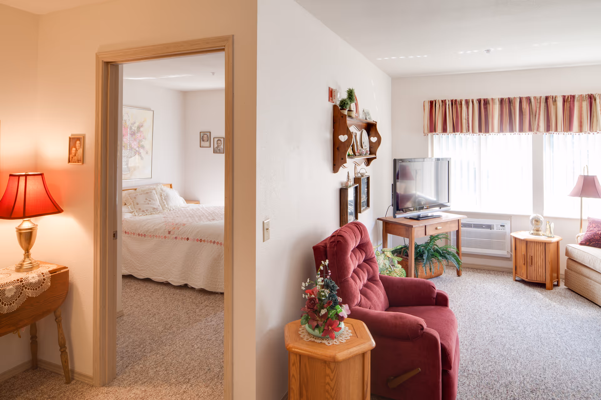 Cozy senior living apartment living room with a red armchair, TV and a view into a bedroom through an open doorway.