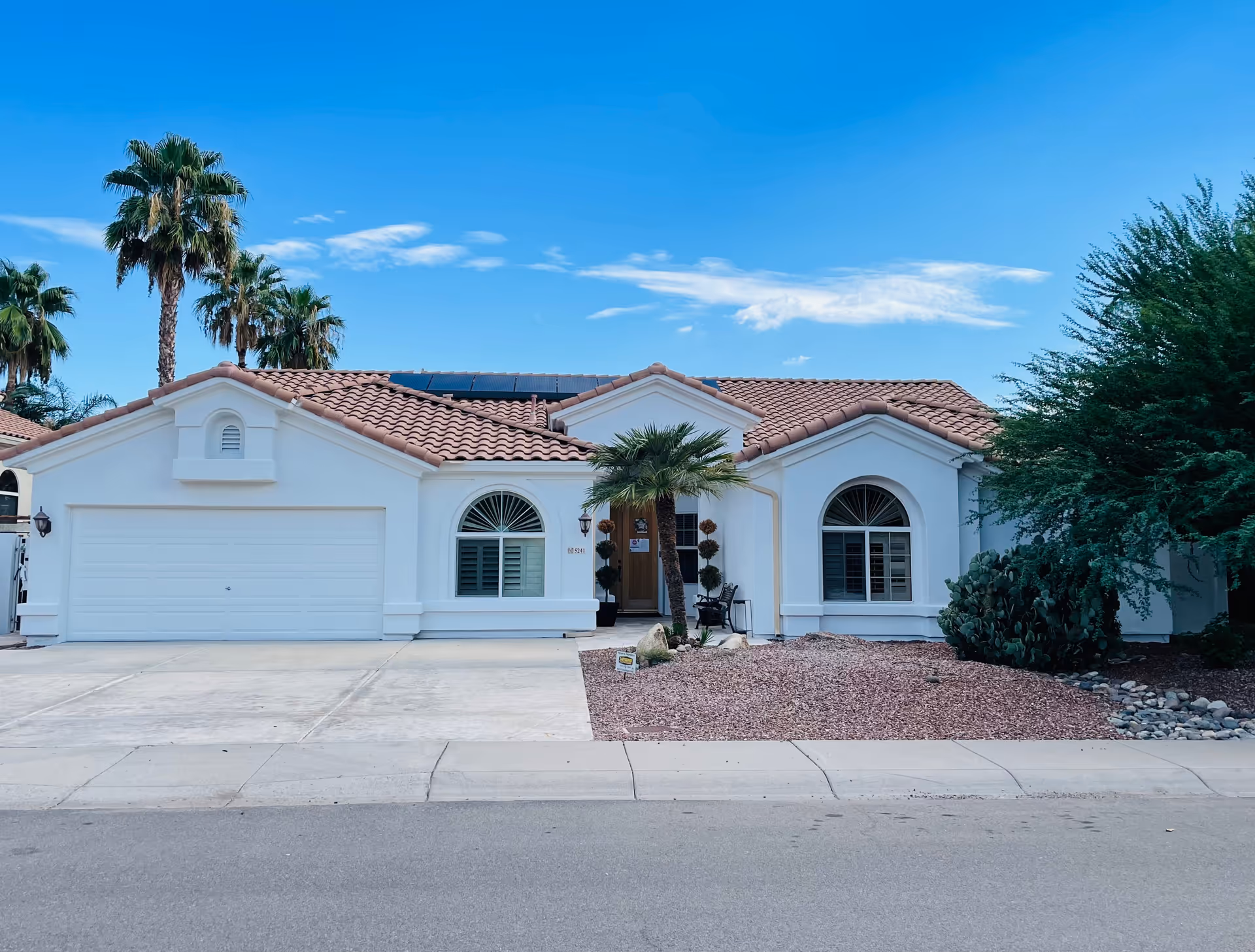 Single-story white stucco house with a red tile roof, attached garage, palm trees, and desert landscaping under a blue sky.