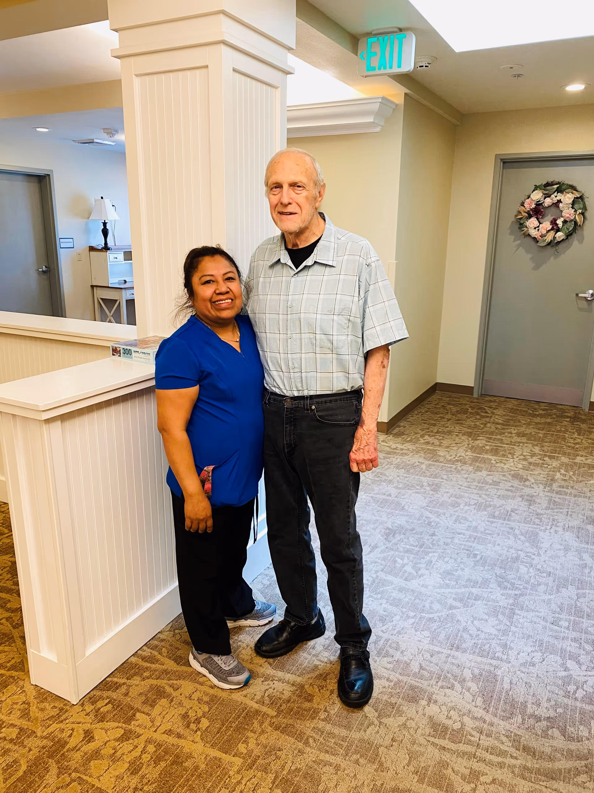 A woman in blue scrubs and a man in a light plaid shirt and black pants stand together smiling in a hallway of a senior living facility. The hallway has beige walls, a carpeted floor, a white column, a door decorated with a floral wreath, and an illuminated exit sign above.
