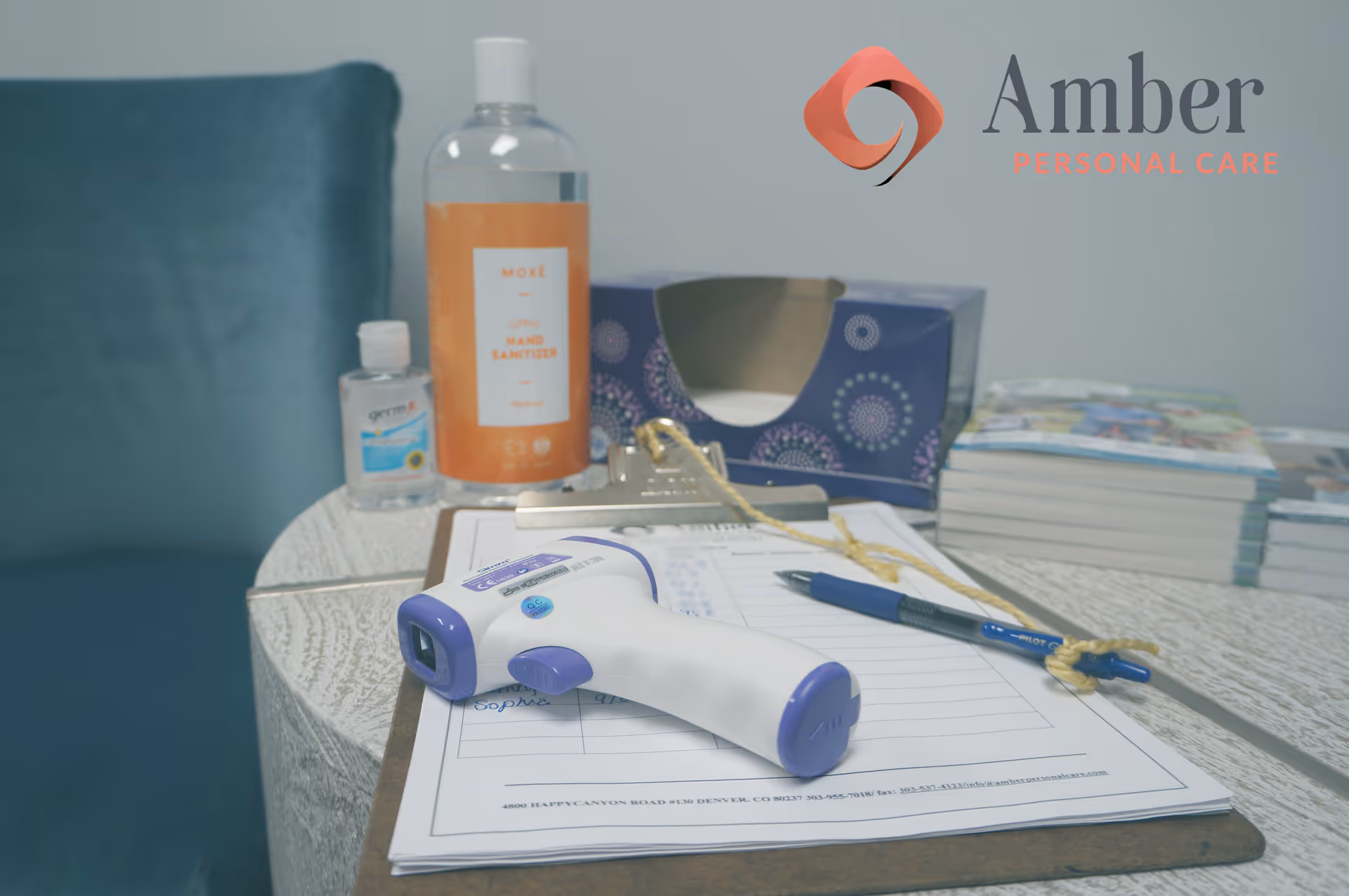 A desk with sanitizer, a thermometer, paperwork, and Amber Personal Care branding.