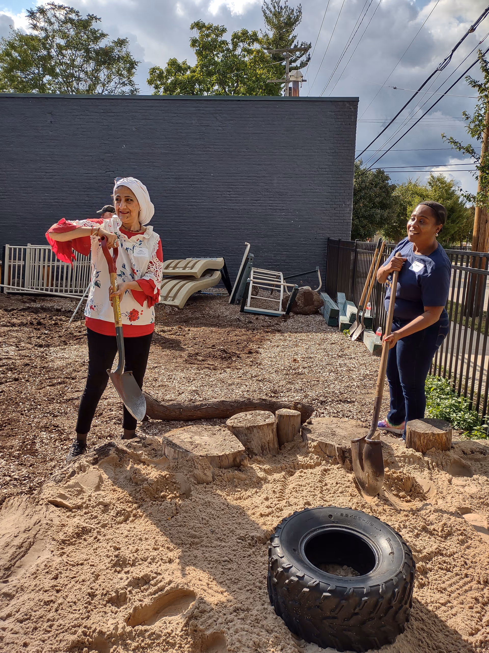 Two women shovel sand in a fenced outdoor play area near a dark building.