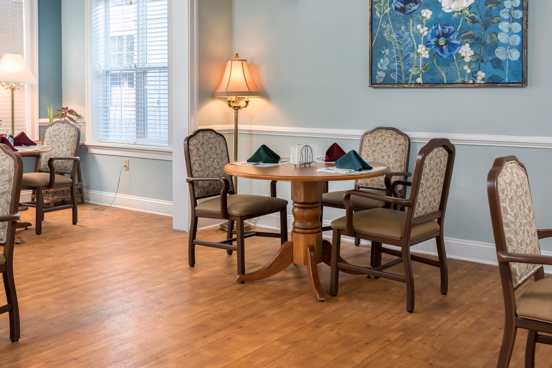 Bright dining room with round wooden table, upholstered chairs, and folded napkins.