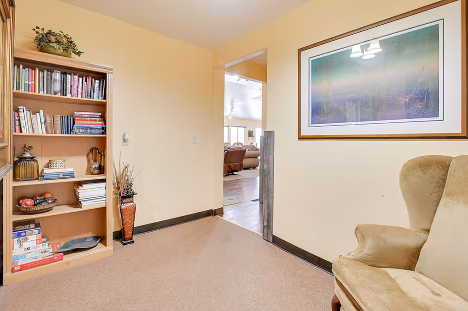 A cozy interior corner of a senior living facility with a wooden bookshelf filled with books and decorative items on the left, a beige armchair on the right, and a framed abstract artwork hanging on the wall. The room has light yellow walls and a carpeted floor. An open doorway leads to a larger common area with more seating visible in the background.