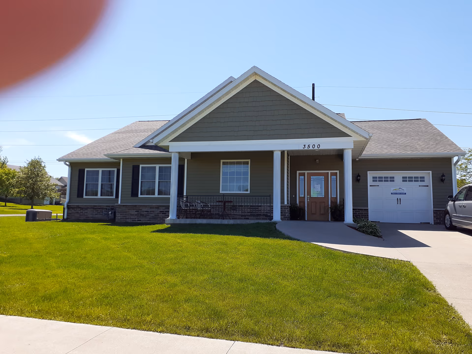 Single-story building with a green exterior, white trim, and a front porch supported by two white columns. The building has a garage on the right side and a driveway with a parked vehicle. The lawn in front is well-maintained and green under a clear blue sky. The address number 3500 is displayed above the porch.
