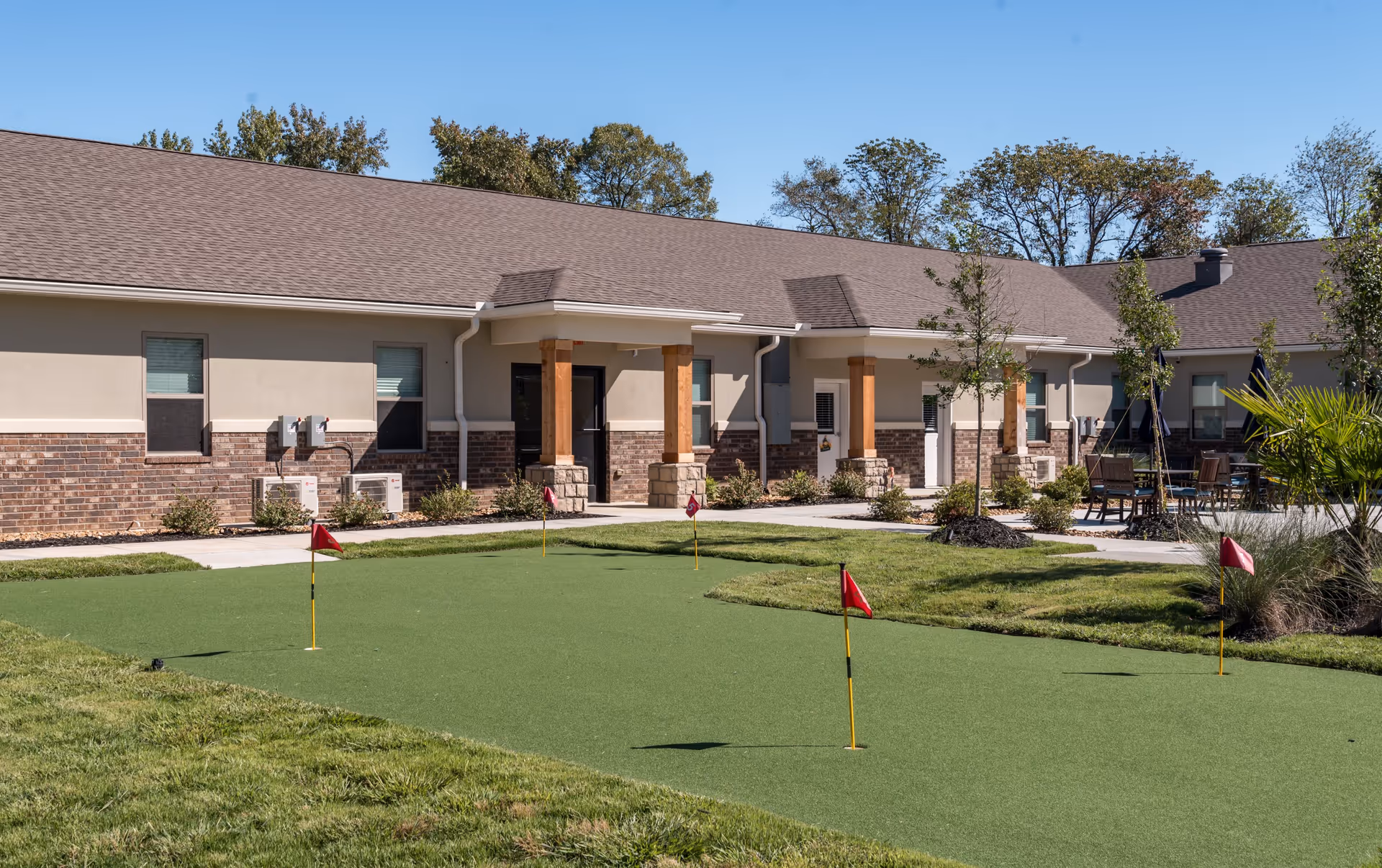 Exterior view of a single-story senior living building with a putting green and small flags in the foreground.