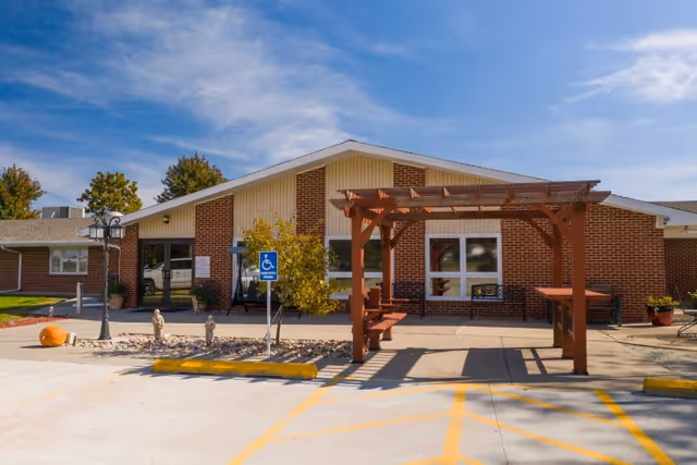 Front exterior of a single-story brick nursing and rehabilitation center with a wooden pergola, benches, and a handicapped parking sign.