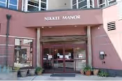 Front entrance of Nikkei Manor with glass doors, signage, and potted plants along a brick facade.