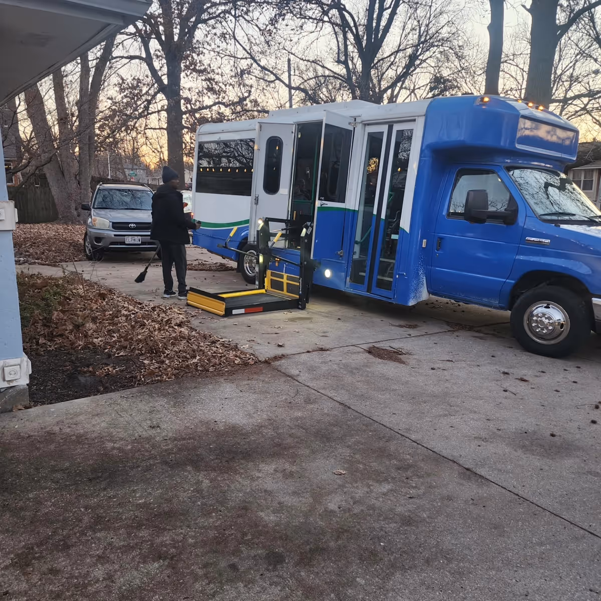 Blue transit bus with a ramp at a driveway; person sweeping leaves nearby.