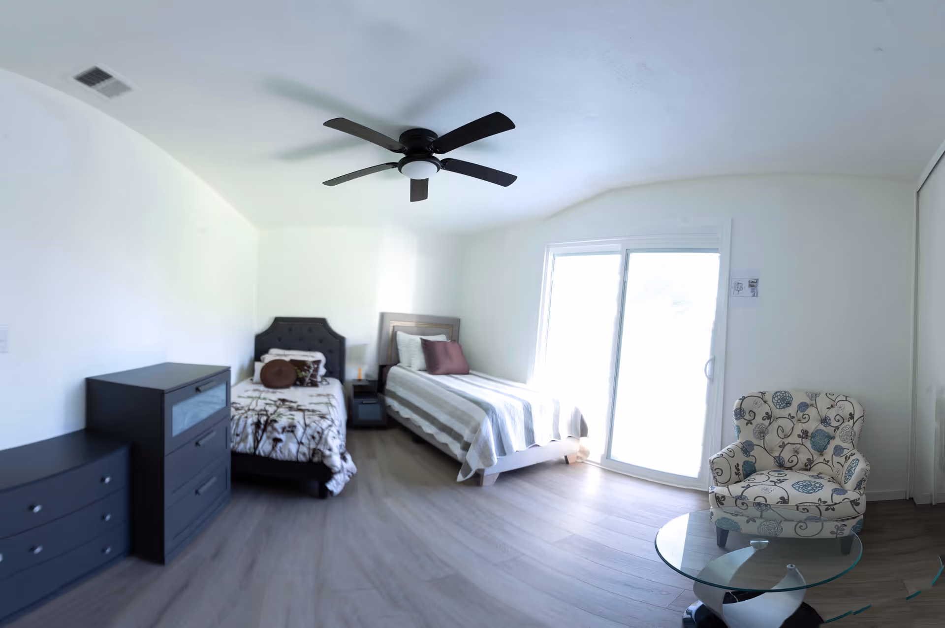 Bright bedroom with two beds, dressers, chair, and sliding glass door in a care facility.