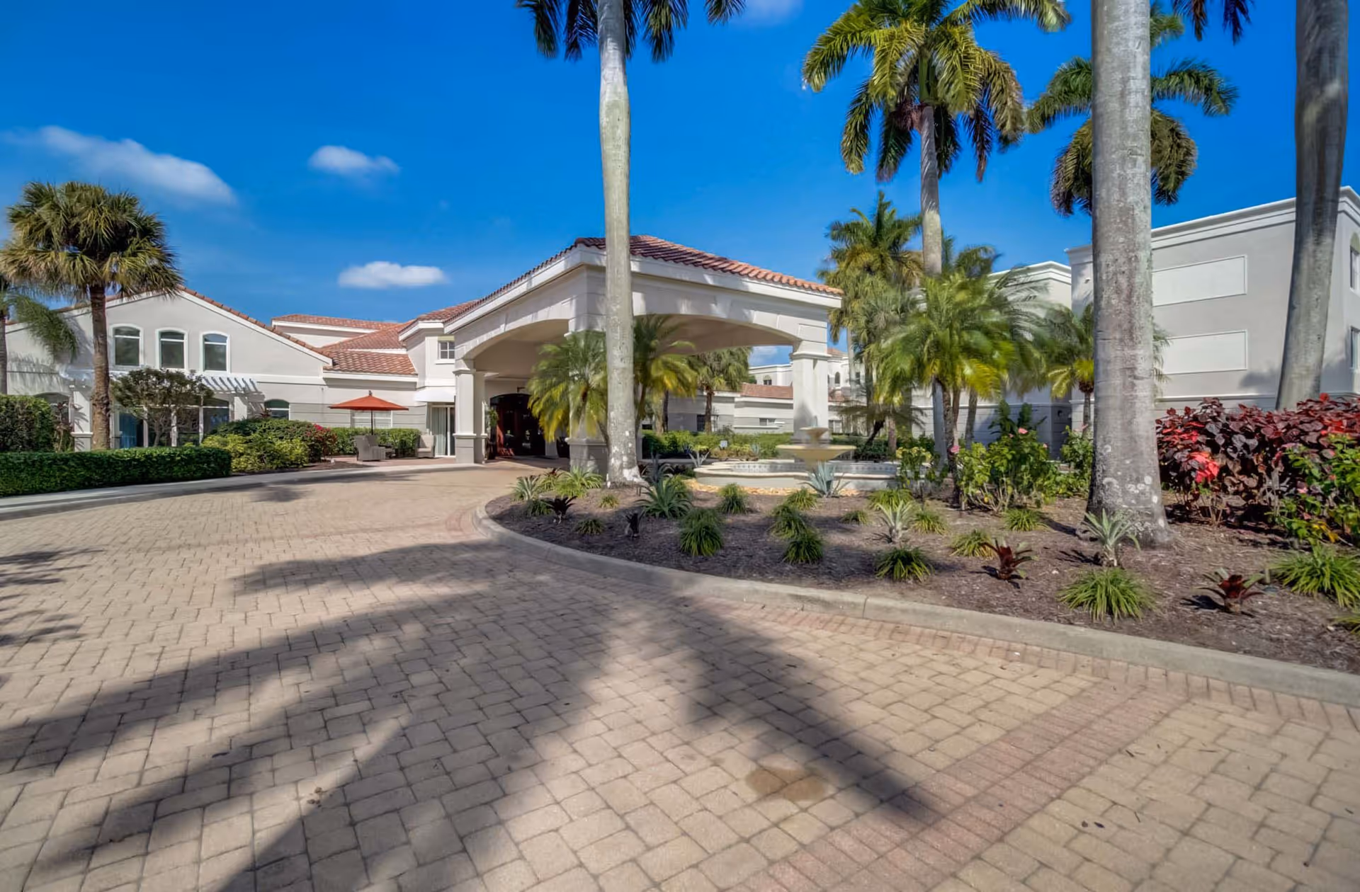 Exterior view of The Carlisle Naples facility showing a paved driveway leading to a covered entrance with columns. The building has a light-colored facade with red-tiled roofing. There are palm trees and landscaped plants around the entrance under a clear blue sky.