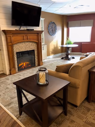 A cozy living room with a couch and side table in front of an electric fireplace topped by a wall-mounted TV, with a round table and window in the background.