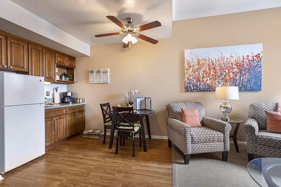 A cozy interior space featuring a small kitchen area with wooden cabinets, a white refrigerator, and a coffee maker on the counter. Adjacent to the kitchen is a round dining table with four chairs. Next to the dining area are two patterned armchairs with pink cushions, a small side table with a lamp, and a colorful abstract painting on the wall. The room has wood flooring and a ceiling fan with lights.