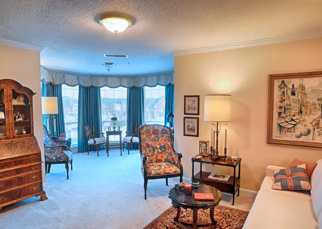 Bright, traditionally furnished living room with floral armchairs, a sofa, coffee table, side table with lamp, a wooden display cabinet, and a bay window with teal curtains.