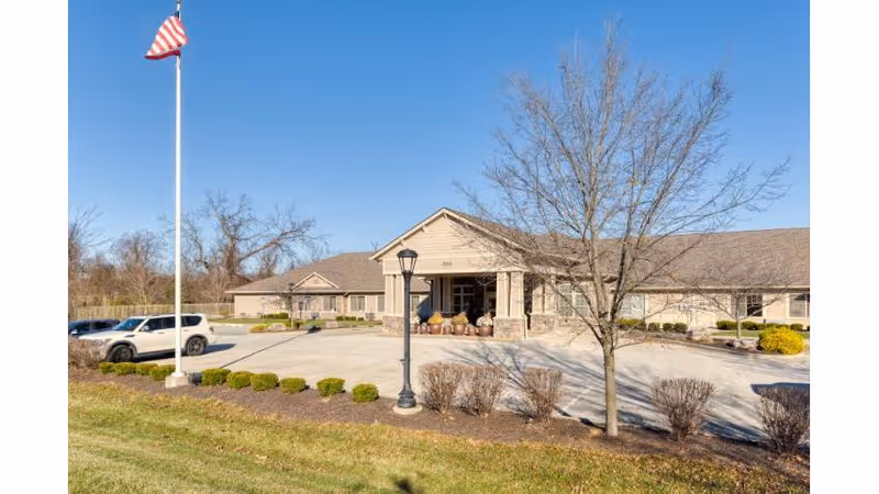 Exterior view of Cedar Creek of Bloomington Memory Care facility on a clear day. The building is a single-story structure with a covered entrance supported by columns. There is a flagpole with an American flag and a parking area with a few cars. Leafless trees and trimmed bushes are visible in the landscaped area in front of the building.