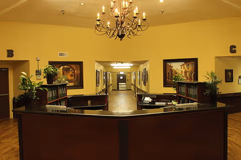 A warmly lit senior living lobby with a curved reception desk and long hallway.