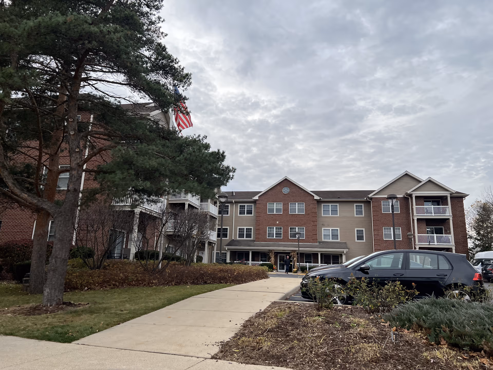 Exterior view of a three-story senior living facility building with a mix of brick and siding. There is a sidewalk leading to the entrance, a large tree on the left, some bushes, and a parked black car on the right. The sky is cloudy.