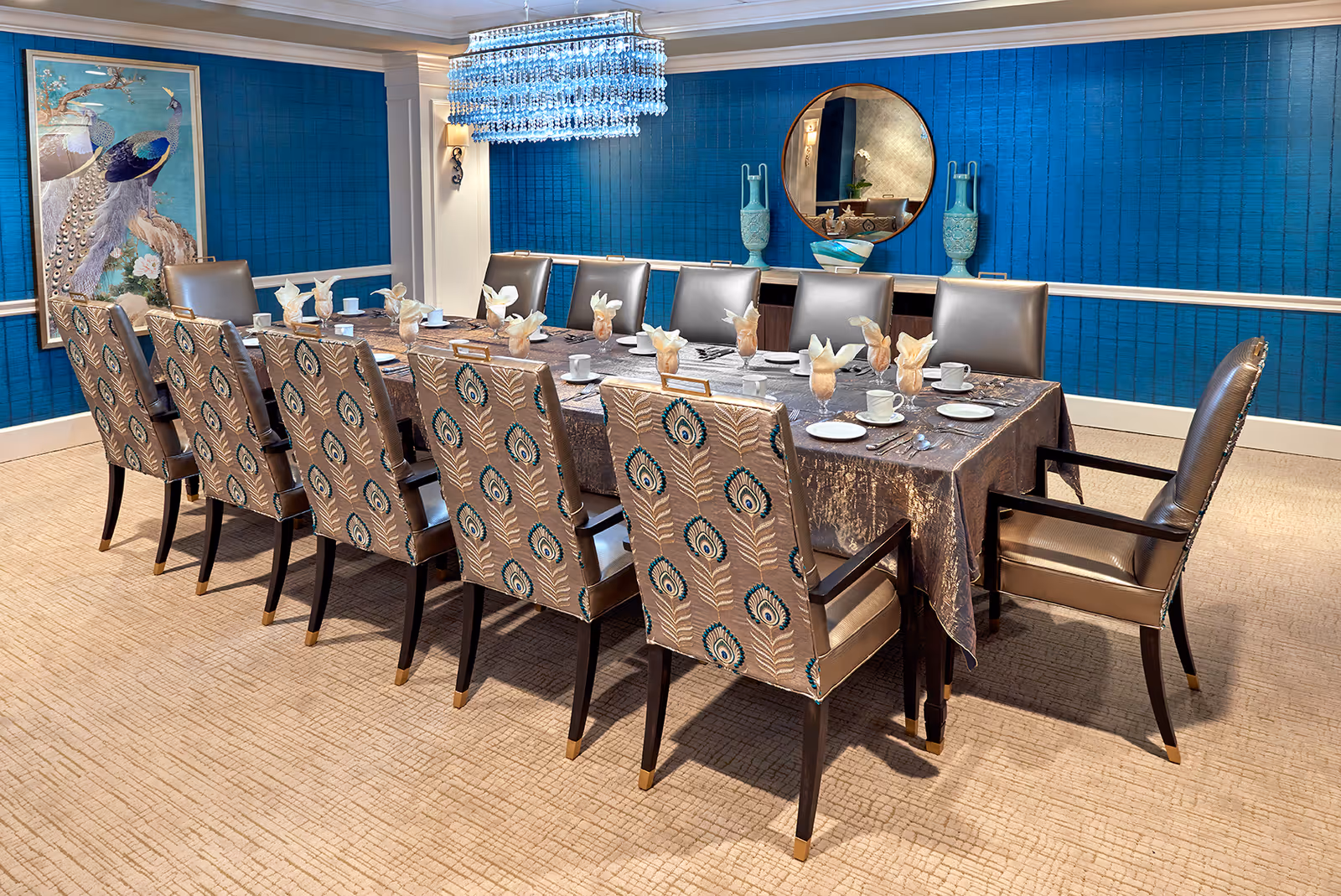A formal dining room with a long rectangular table covered with a dark tablecloth, set with white cups, plates, and napkins folded in an elegant style. The table is surrounded by twelve chairs, six with peacock feather patterned upholstery and six with plain leather upholstery. The room features a blue textured wall with a large round mirror and decorative vases, a framed peacock artwork, and a modern chandelier hanging above the table.