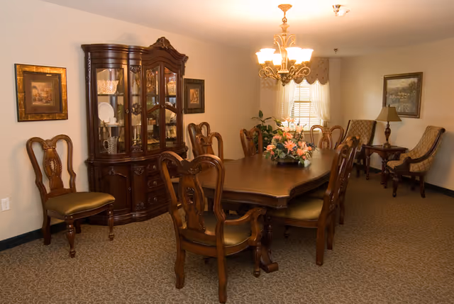 A traditional dining room with a large wooden dining table surrounded by eight matching wooden chairs with upholstered seats. A floral centerpiece is on the table. Against the wall is a wooden china cabinet displaying glassware and dishes. The room is softly lit by a chandelier hanging above the table. There are framed paintings on the walls, a window with sheer curtains, and two upholstered armchairs with a small side table and lamp in the corner.