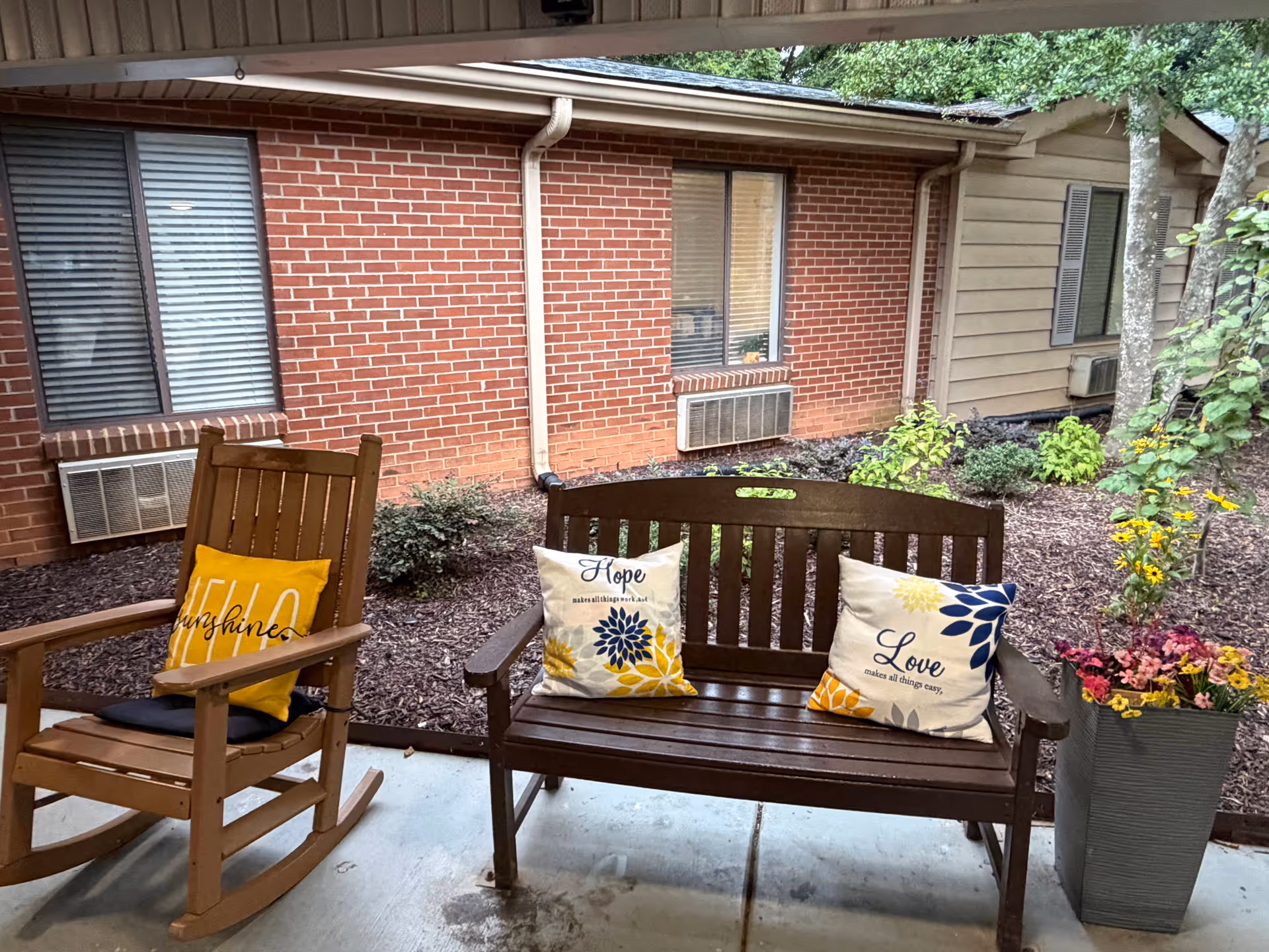 Wooden bench and rocking chair with cushions on a covered outdoor patio beside a brick building.