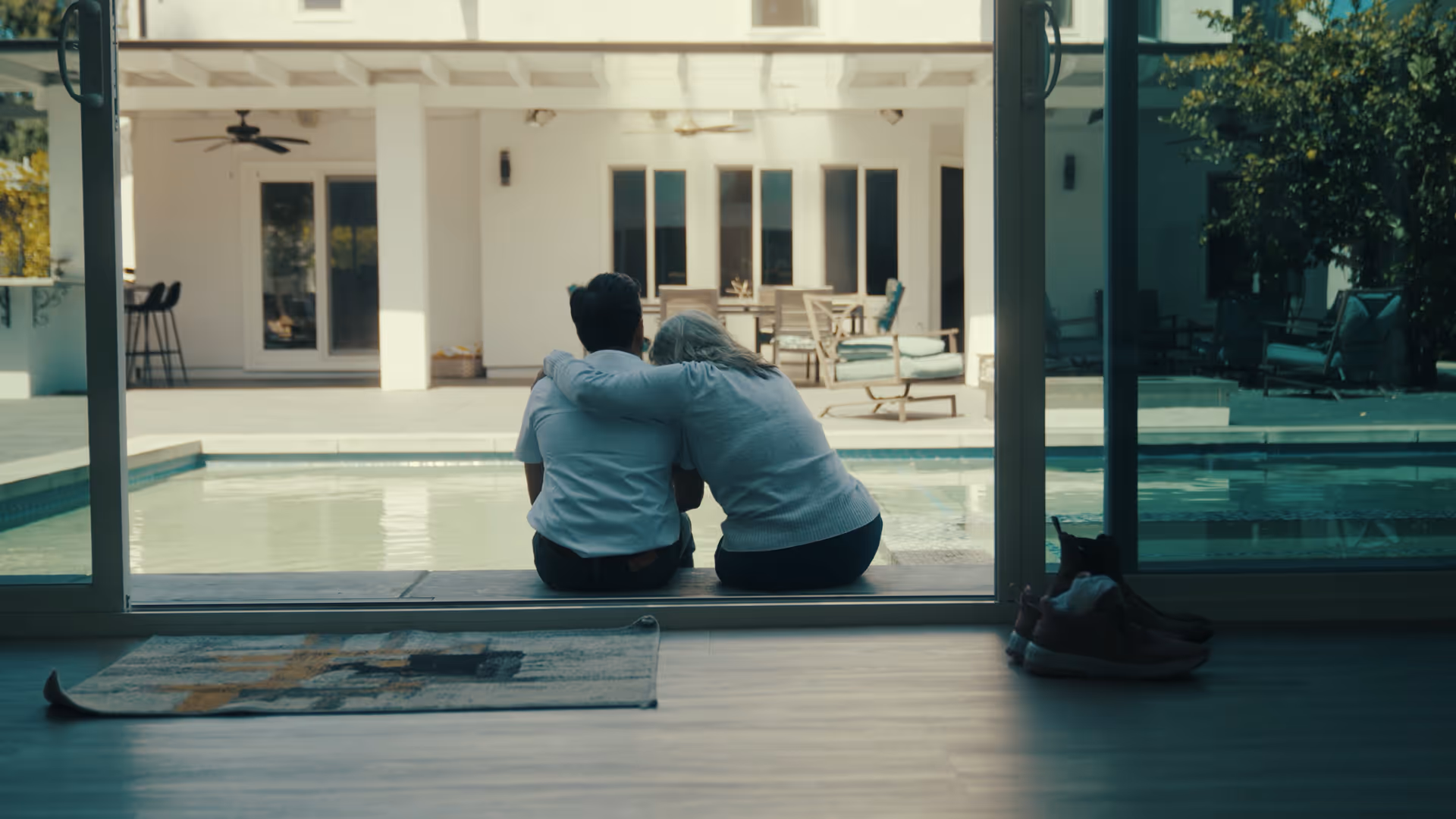 Two people sit close together on a sliding-glass door threshold, overlooking a poolside backyard.