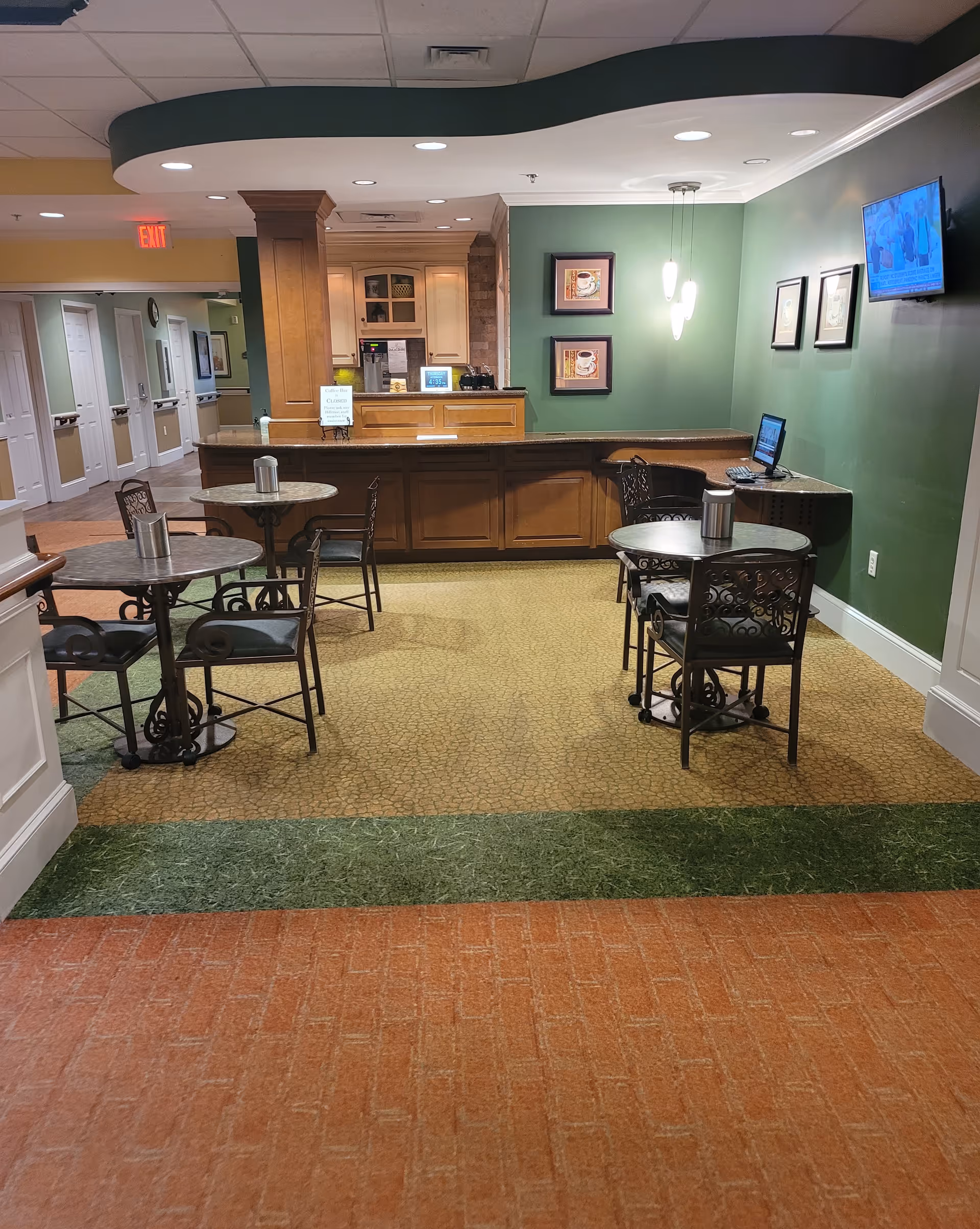 Reception lobby with wooden front desk, green walls, round tables, chairs, and a wall TV