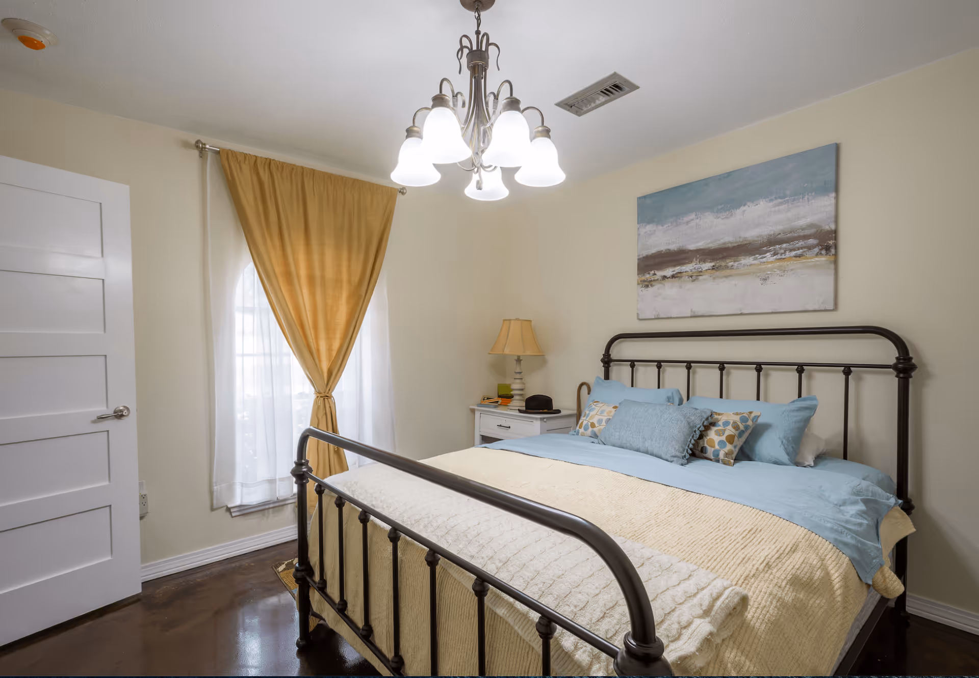 Cozy bedroom featuring a metal bed frame, nightstand, lamp, beige curtains, and blue bedding.