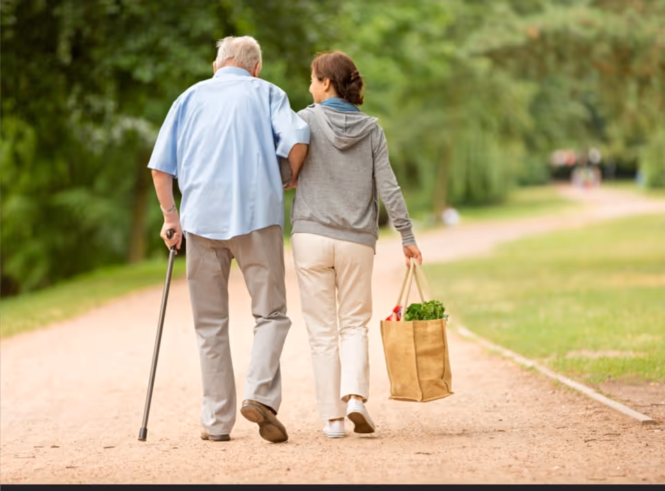 An elderly couple walks along a park path, one using a cane, carrying a shopping bag.