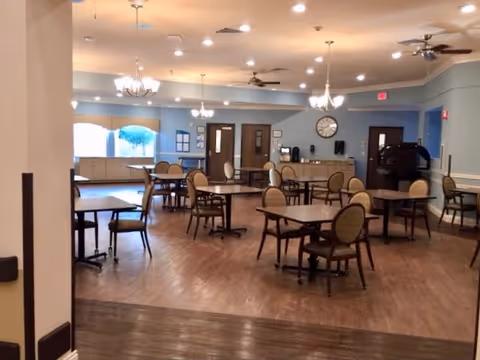 A spacious dining room with multiple square tables and chairs arranged neatly on a wooden floor. The room has light blue walls, several ceiling fans, and chandeliers providing warm lighting. There is a clock on the far wall above a counter area with coffee machines and other appliances. Large windows allow natural light to enter the room.