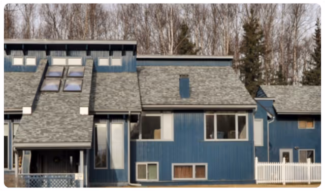 Blue multi-unit building exterior with pitched shingle roofs, skylights, windows, and a white picket fence against a backdrop of leafless trees.