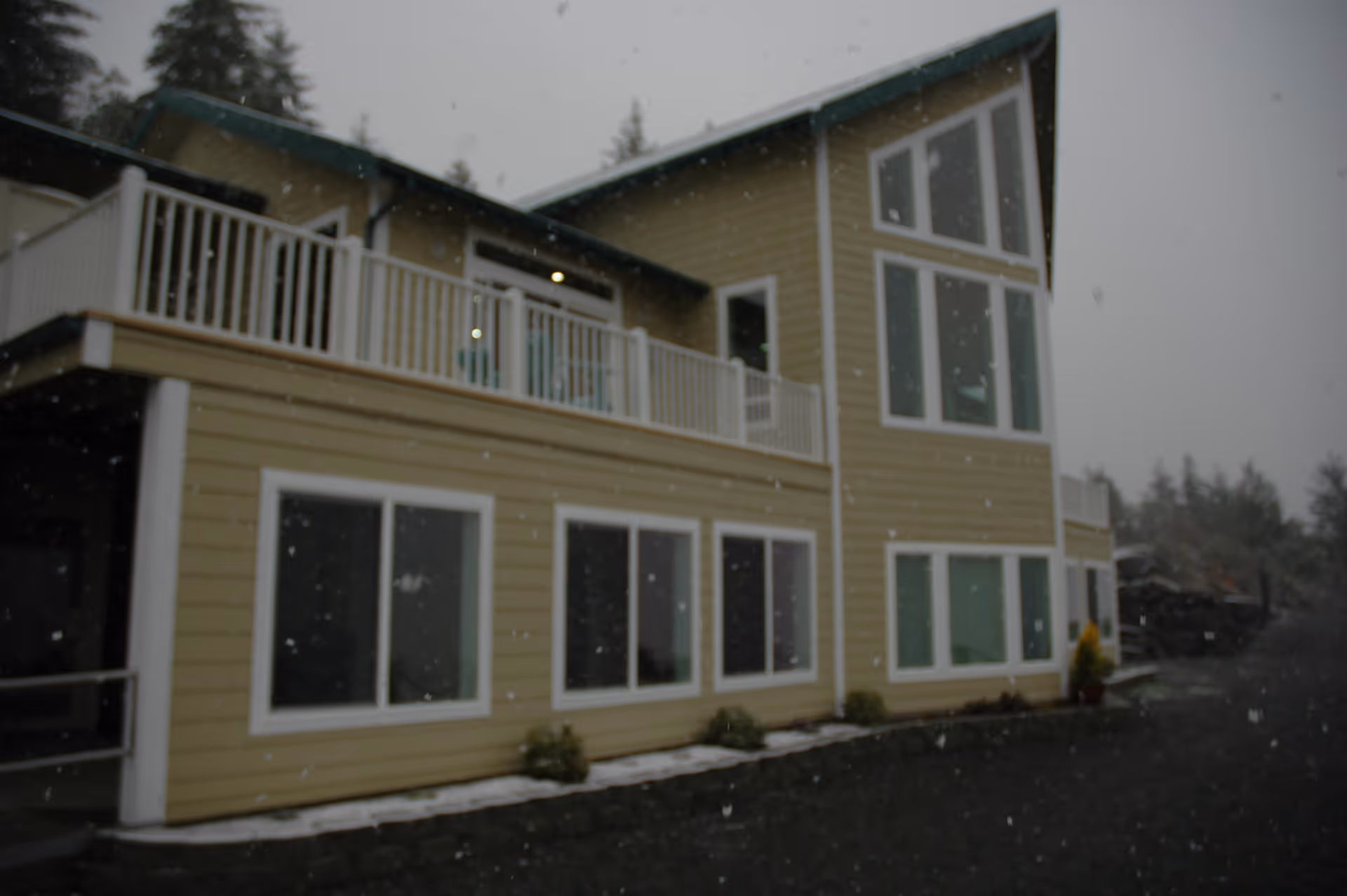 Exterior view of a two-story yellow building with white trim and large windows, featuring a balcony with white railings. Snow is falling and some snow is visible on the ground and roof edges. Trees are visible in the background under a gray sky.