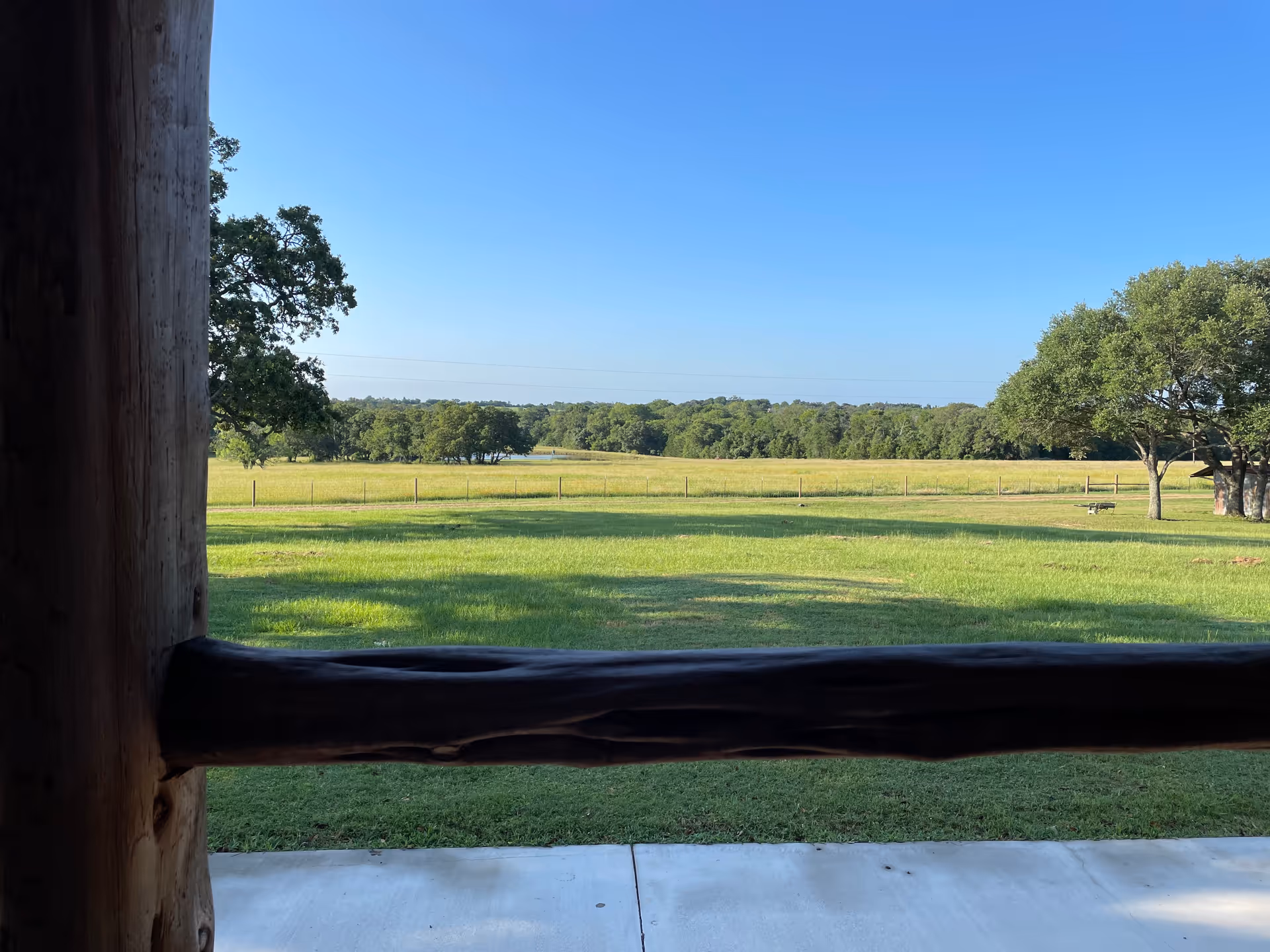 View of a grassy field with trees beyond a wooden railing on a sunny day.