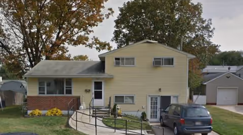 Two-story yellow assisted living building with ramp, front entrance, and a parked car.