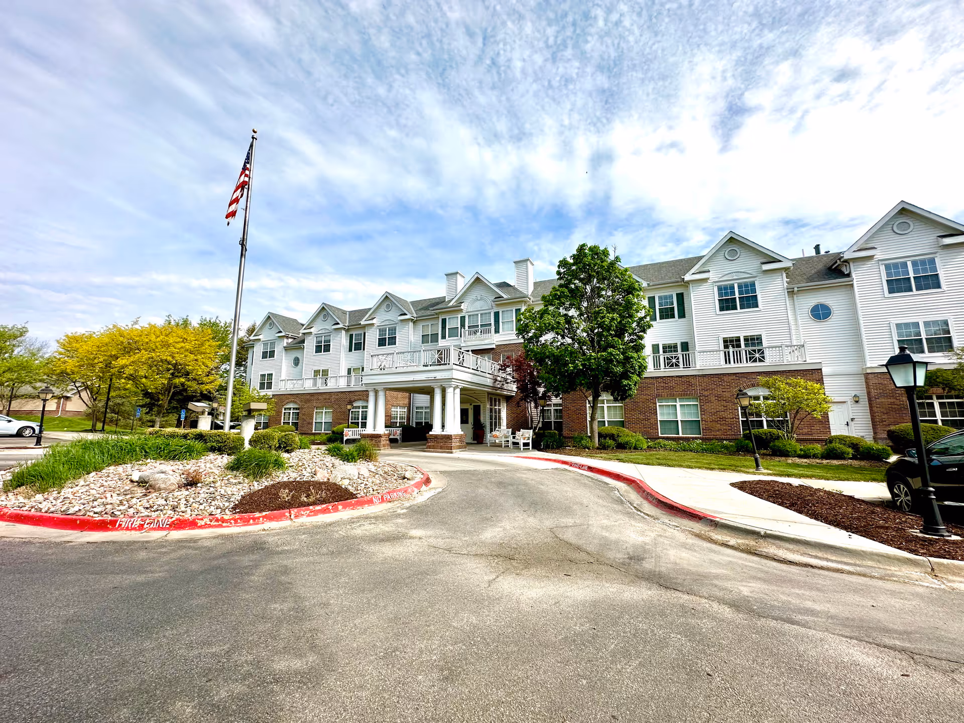 Front exterior view of a senior living facility building with white siding and brick accents, a covered entrance with columns, an American flag on a flagpole, trees, and a driveway with red curbs marked 'FIRE LANE NO PARKING'.