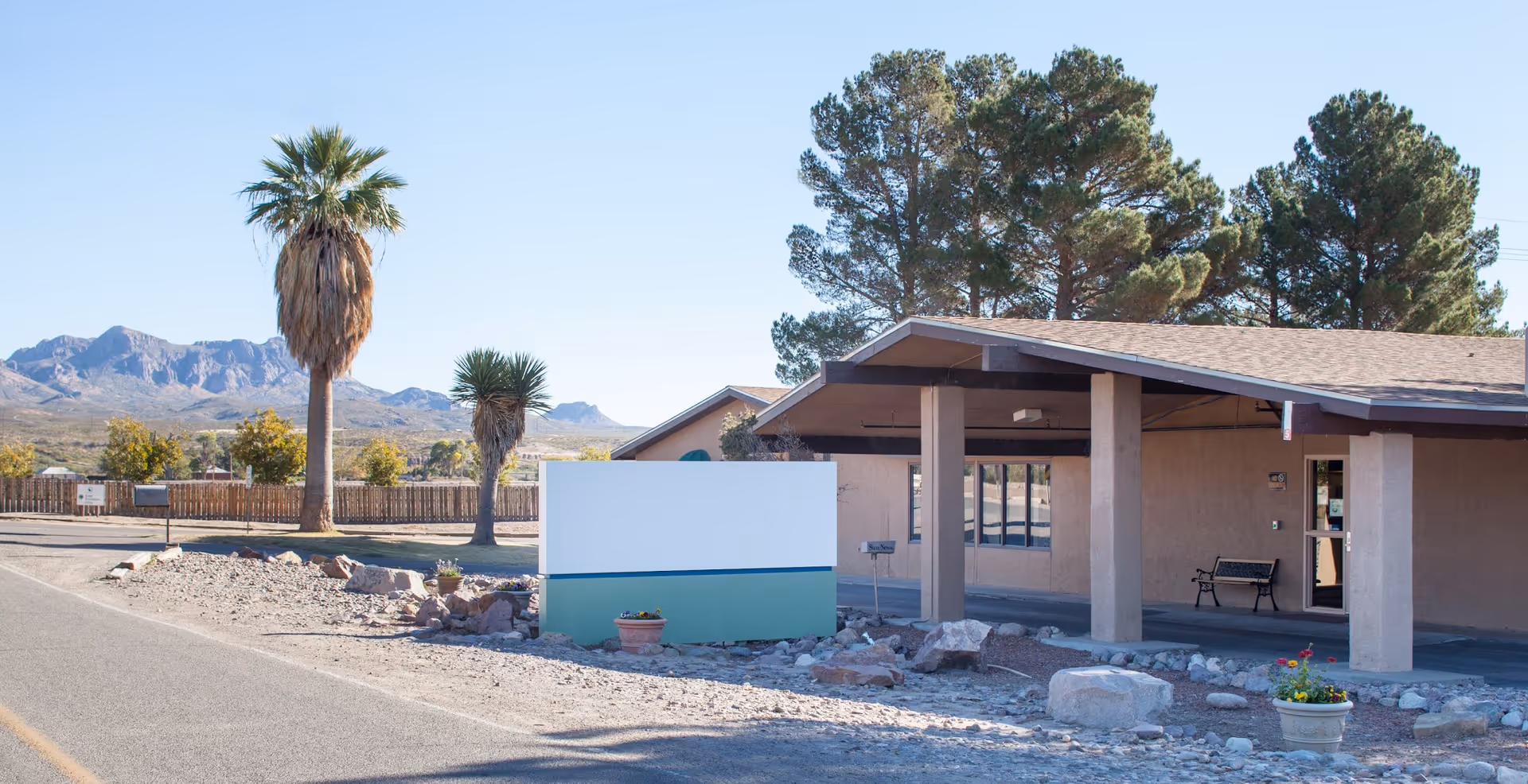 Single-story building exterior with a covered entrance, a blank sign, desert landscaping, palm trees, and distant mountains.